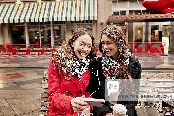 Glückliche Freundinnen telefonieren  während sie in der Stadt auf einer Bank sitzen