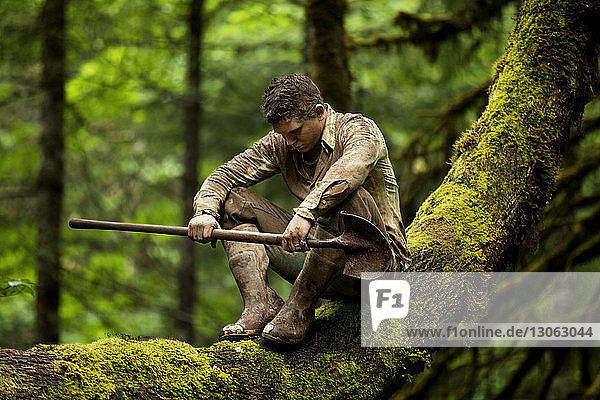 Bauer sitzt auf Baumstamm und hält Schaufel im Wald