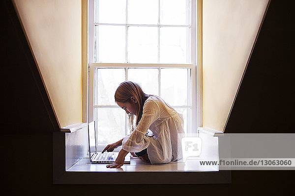 Mädchen benutzt Laptop-Computer  während sie zu Hause am Fenster sitzt