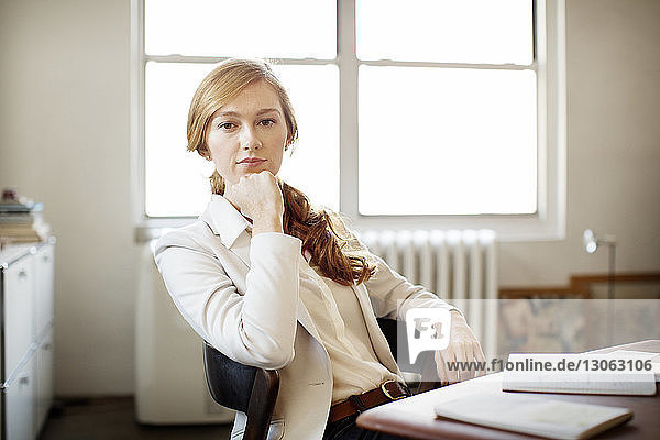 Portrait of confident businesswoman with hand on chin sitting at desk in office