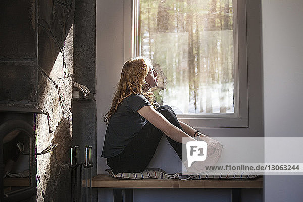 Nachdenkliche Frau schaut durch das Fenster  während sie zu Hause auf der Bank sitzt