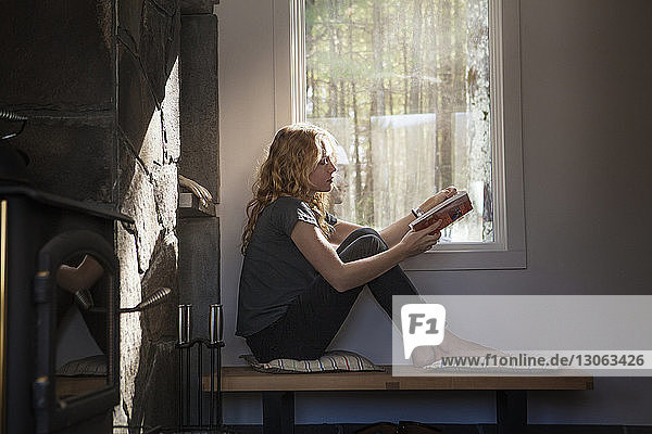Frau liest Buch  während sie zu Hause am Fenster sitzt
