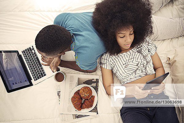 Couple having croissants and using technology while resting on bed at home