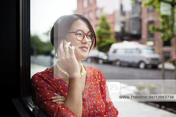 Happy woman talking on smart phone while standing by window