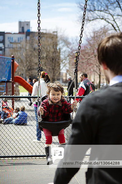 Glückliches Mädchen sitzt auf der Schaukel  während der Vater auf dem Spielplatz steht