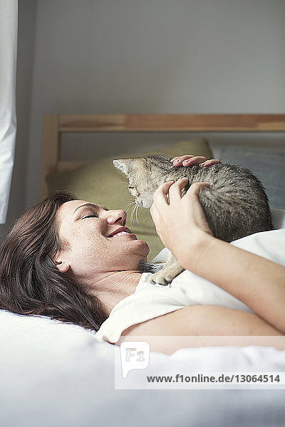 Lächelnde Frau spielt mit Katze  während sie zu Hause auf dem Bett liegt