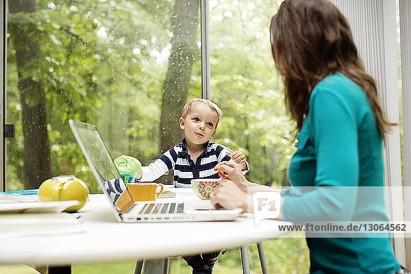 Mutter unterhält sich mit ihrem Sohn  während sie am Tisch einen Laptop benutzt