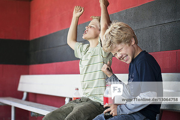 Brothers enjoying while sitting on bench at baseball dugout