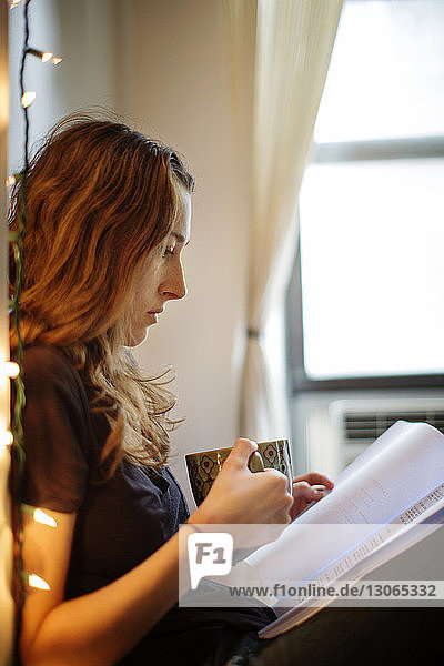 Side view of woman holding coffee cup while reading book at home