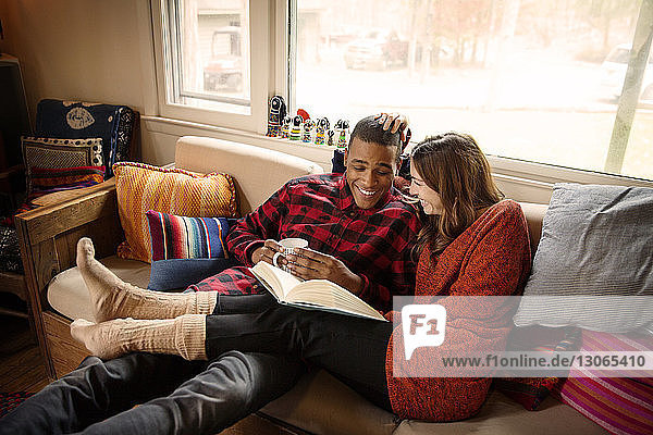 Ein Paar liest ein Buch  während es zu Hause auf dem Sofa Kaffee trinkt