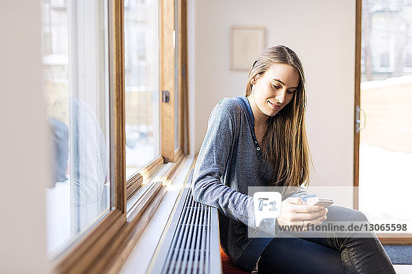 Frau benutzt Smartphone  während sie zu Hause am Fenster sitzt