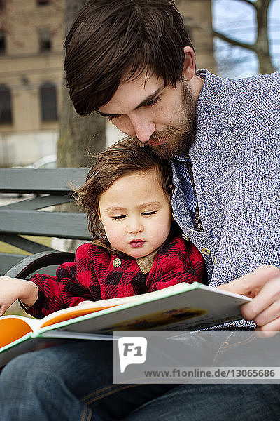 Mann liest Buch  während er mit seiner Tochter auf der Bank sitzt