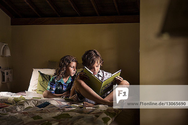 Siblings reading book while sitting on bed in cabin