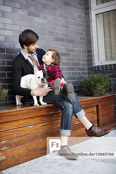Happy father and daughter with dog sitting on bench against wall