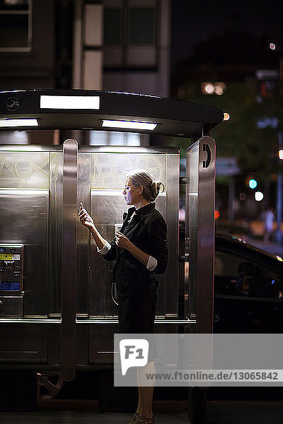 Seitenansicht einer Geschäftsfrau  die telefoniert  während sie an beleuchteten Telefonkabinen in der Stadt steht