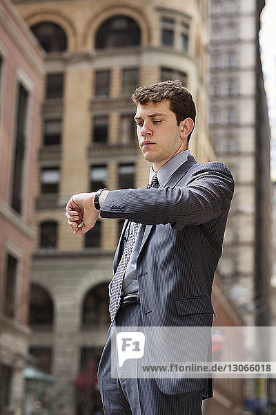 Businessman looking at wristwatch while standing against buildings in city