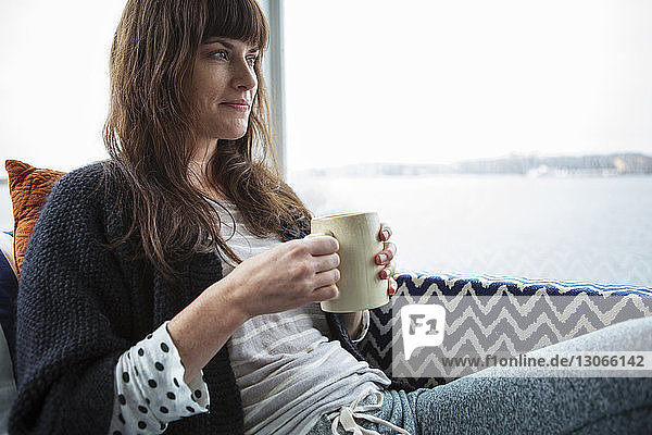 Nachdenklich lächelnde Frau trinkt Kaffee  während sie zu Hause am Glasfenster sitzt