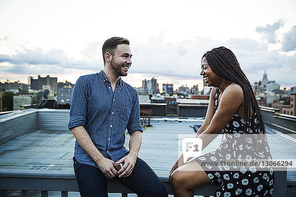Glückliche Freunde unterhalten sich  während sie auf dem Geländer einer Gebäudeterrasse vor bewölktem Himmel sitzen