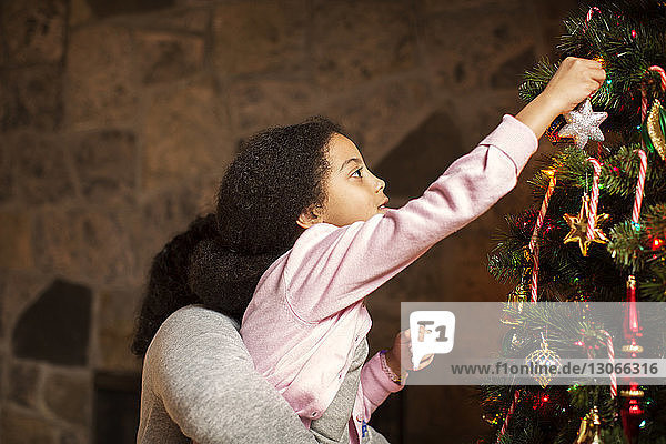 Seitenansicht einer Mutter  die eine Tochter trägt  die zu Hause den Weihnachtsbaum schmückt