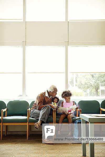 Grandfather looking at girl reading book while sitting on chair in hospital