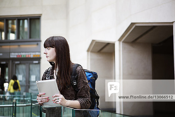 Frau mit Tablet-Computer schaut weg  während sie am Geländer des Bahnhofs steht