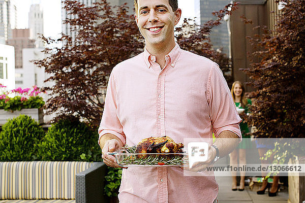 Midsection of smiling man carrying roasted chicken in tray while standing against plants