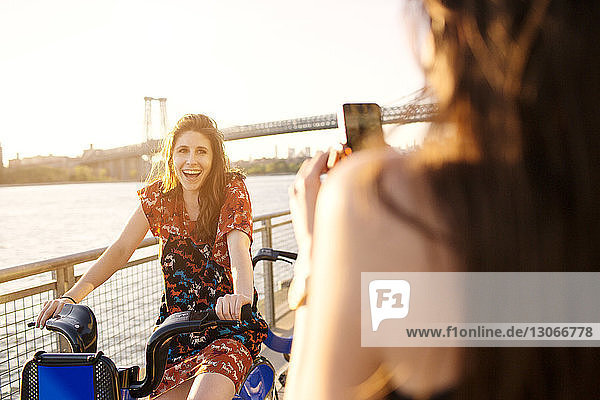 Woman photographing friend on bicycle against clear sky