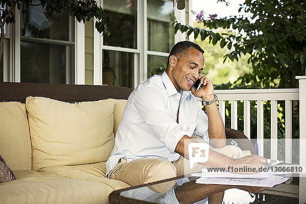 Man talking on mobile phone while writing on paper in porch