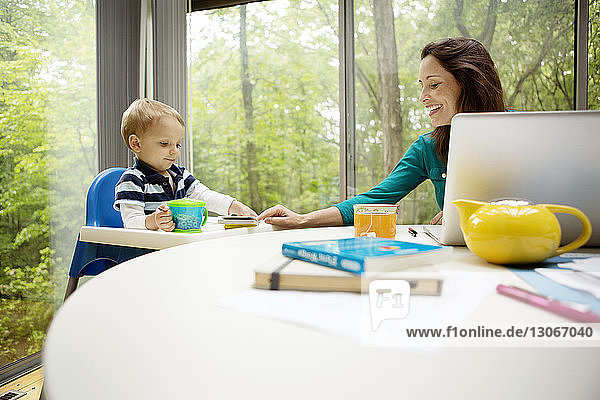 Lächelnde Mutter unterrichtet Sohn  während sie Laptop am Tisch gegen Fenster benutzt