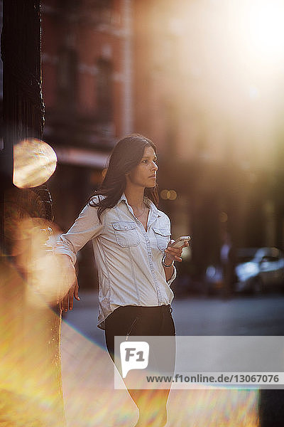 Woman looking away while holding smart phone at street