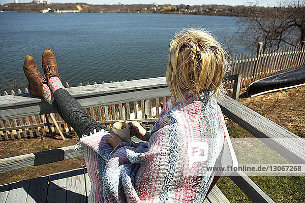 In eine Decke gehüllte Frau trinkt Kaffee  während sie auf einer Bank am See sitzt