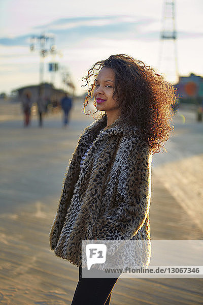Lächelnde Frau mit lockigem Haar trägt Pelzmantel  während sie auf Coney Island steht
