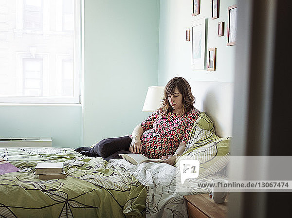 Pregnant woman reading book while sitting on bed