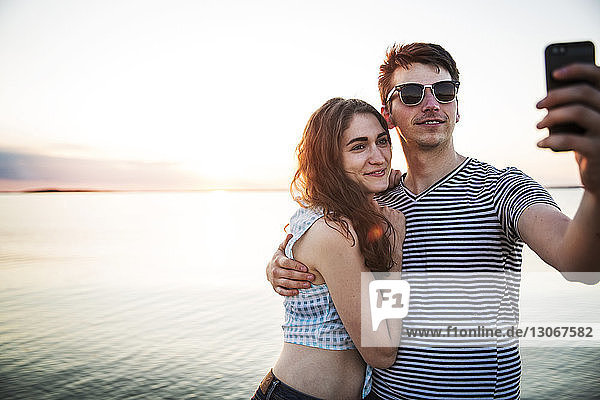 Happy couple taking selfie while standing against sea during sunset