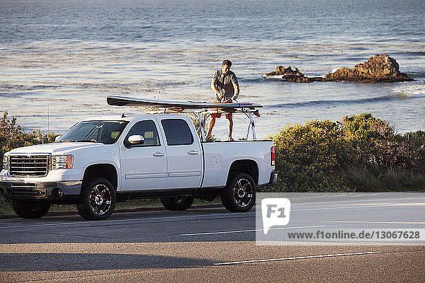 Mann bindet Surfbrett auf am Straßenrand geparkten Kleinlaster gegen das Meer