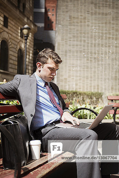 Businessman using laptop computer while sitting on bench against buildings