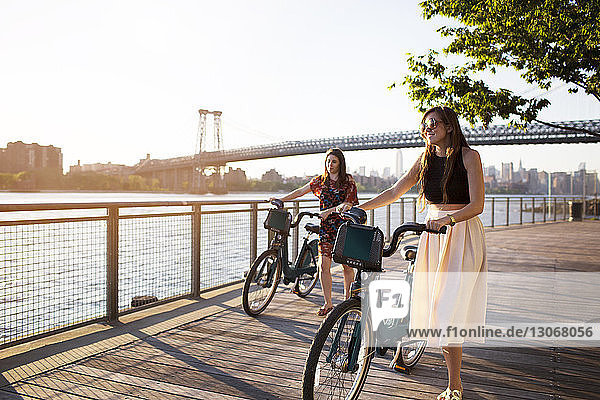 Friends with bicycle walking on promenade against Williamsburg Bridge during sunset