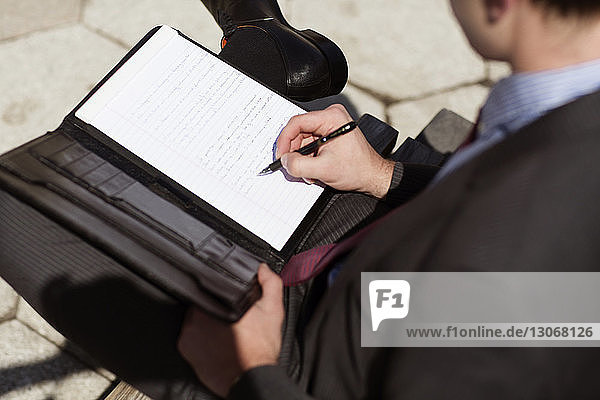 Schrägansicht eines Geschäftsmannes  der auf einem Dokument schreibt  während er auf einer Bank sitzt