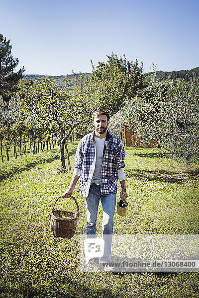 Portrait of man holding basket and wine bottle while standing on field in vineyard
