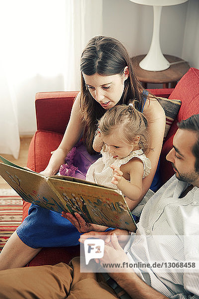 Mann und Frau zeigen einem Mädchen ein Bilderbuch  während sie zu Hause auf dem Sofa sitzen