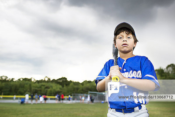 Portrait of boy with baseball bat standing on field against cloudy sky