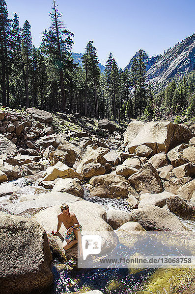 High angle view of shirtless man sitting on rock by stream against clear sky