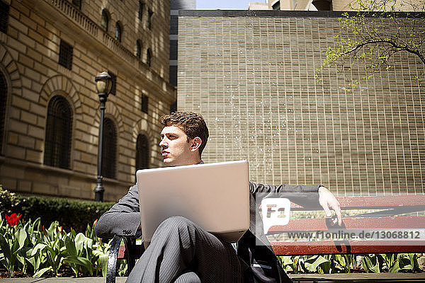 Geschäftsmann mit Laptop-Computer schaut weg  während er auf einer Bank an Gebäuden sitzt