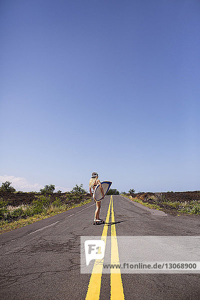 Woman carrying surfboard while skateboarding on road against blue sky