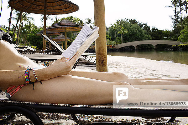 Mittelteil einer Frau  die ein Buch hält  während sie sich auf einem Liegestuhl am Strand entspannt
