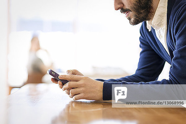 Ausgeschnittenes Bild eines Mannes  der ein Mobiltelefon benutzt  während er sich zu Hause auf einen Tisch lehnt