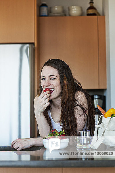 Portrait of woman eating strawberry while leaning on kitchen counter at home