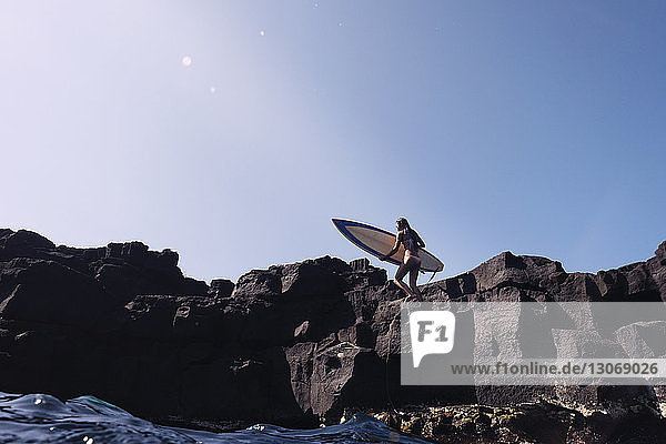 Frau trägt Surfbrett  während sie auf Felsen gegen den klaren Himmel läuft