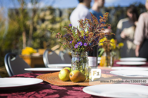 Flower vase and fruits on outdoor table with friends in background at garden party