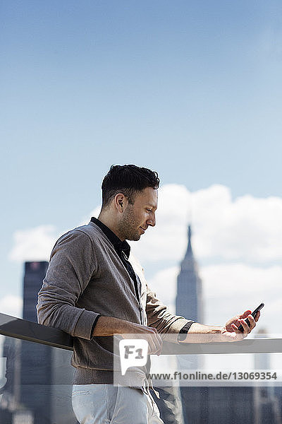 Niedrigwinkelansicht eines Mannes  der ein Mobiltelefon benutzt  während er im Büro auf einem Balkon gegen den Himmel steht